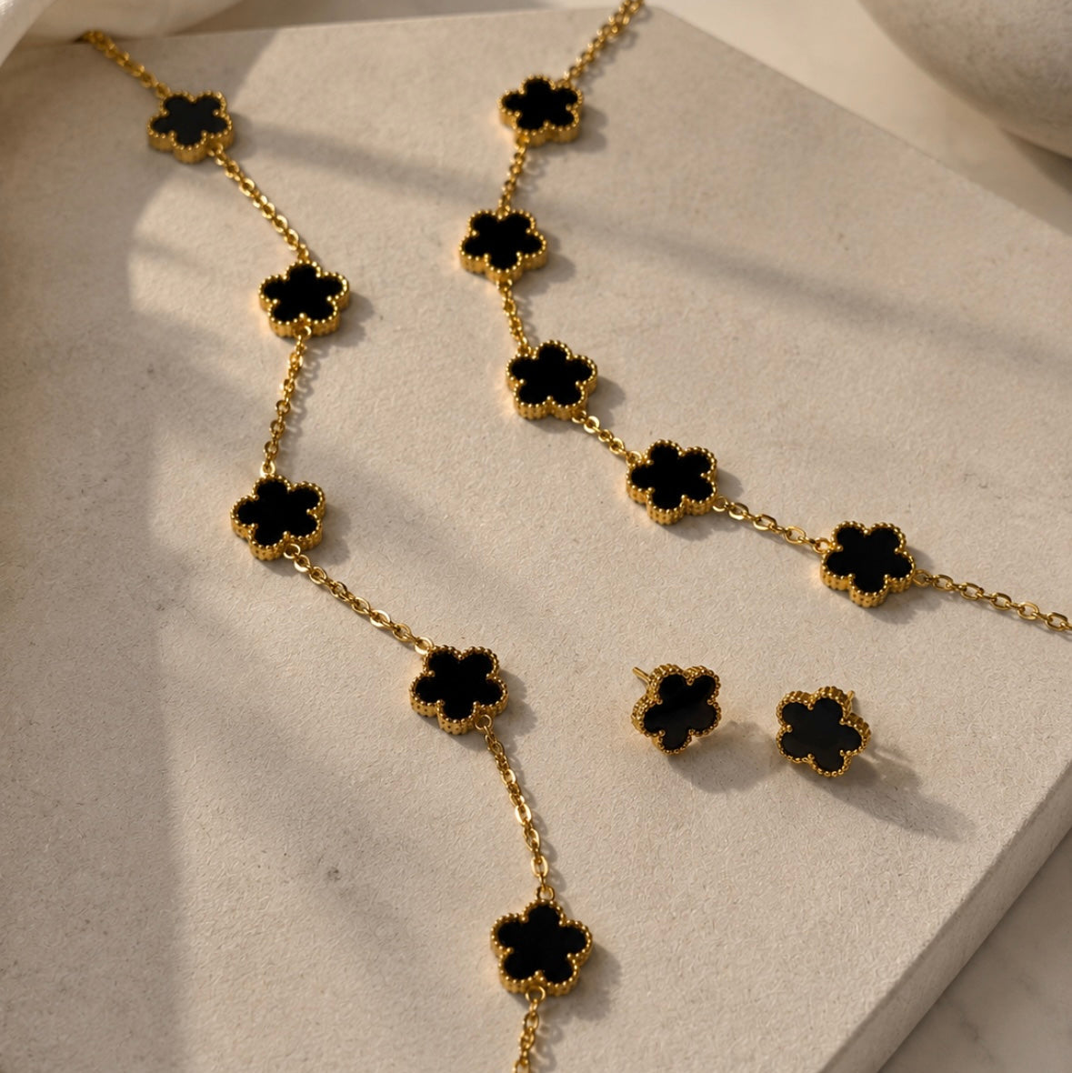 Gold-tone jewelry set with black flower-shaped links, including a bracelet, matching earrings, and a long chain, displayed on a light stone hexagonal tray.