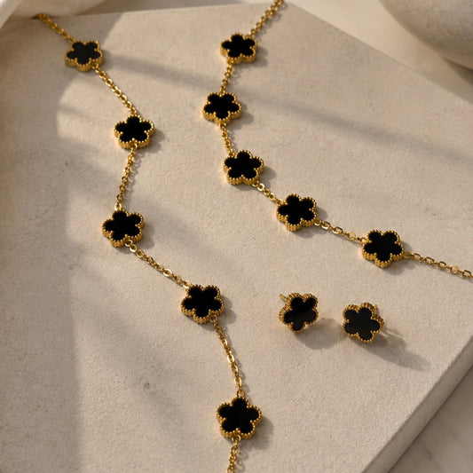 Gold-tone jewelry set with black flower-shaped links, including a bracelet, matching earrings, and a long chain, displayed on a light stone hexagonal tray.